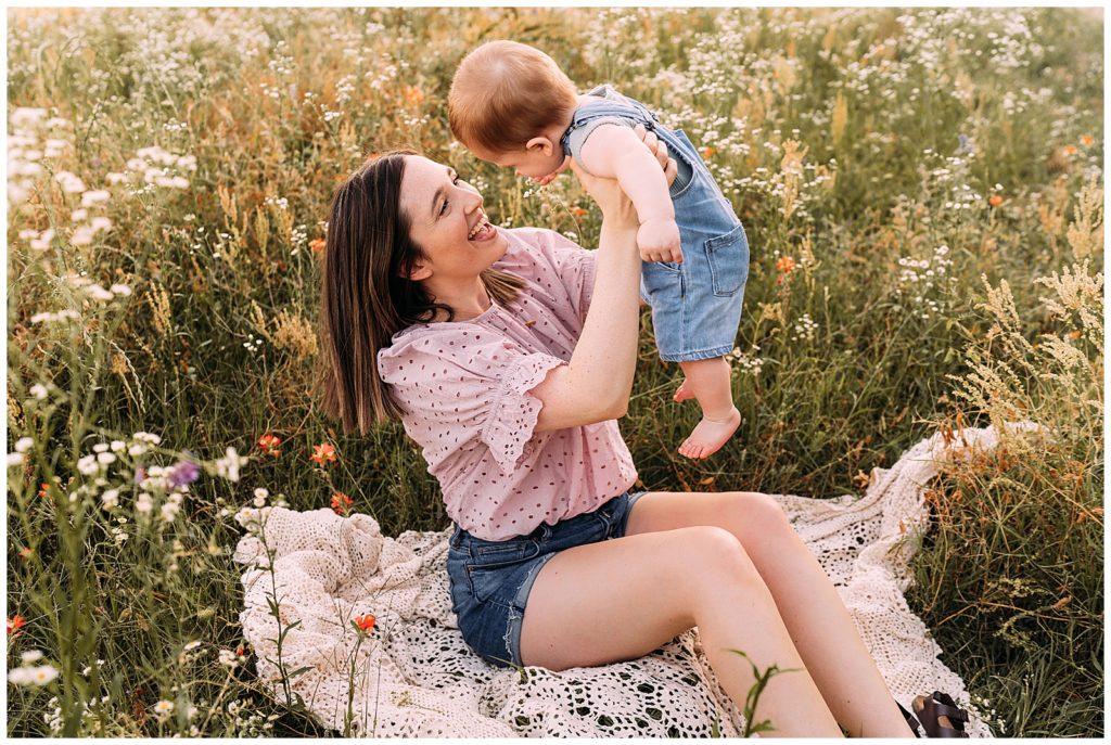 mom lifts 9 month old son into air while sitting in a wildflower field for a lifestyle photography session