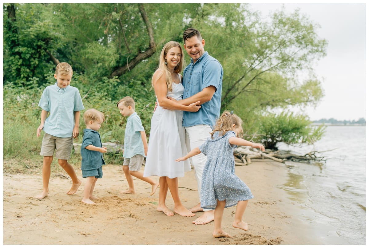 Parents smiling and embracing while children play around them on a sandy beach, an example of what is lifestyle photography with relaxed prompts and authentic connection.