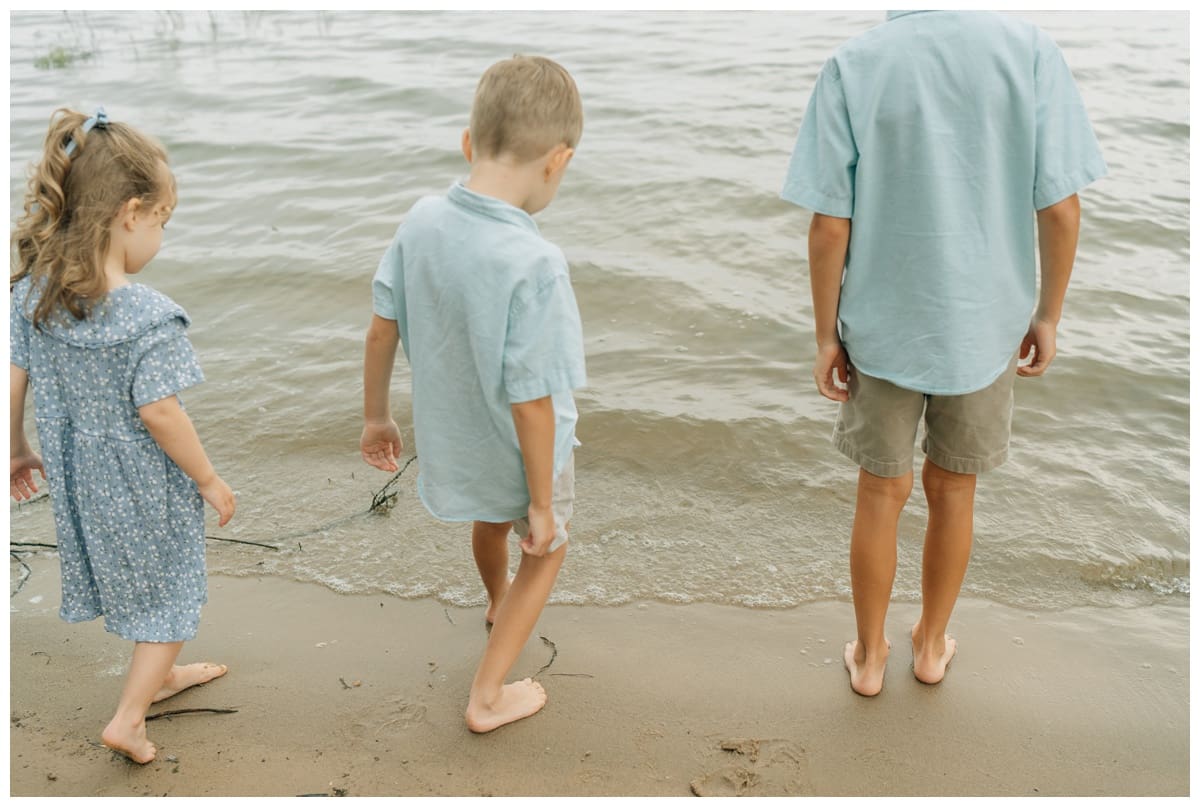 Three young children standing barefoot at the edge of the lake, looking down at the water in Haley Kinzie Photography session.