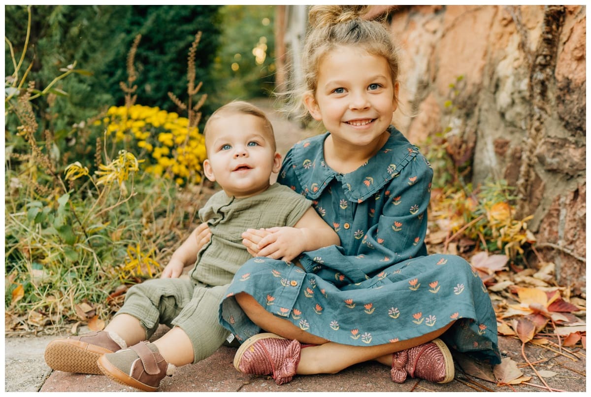Big sister in a teal floral dress hugs her baby brother in a green romper, both smiling while sitting among fall leaves and flowers during a family photography session.