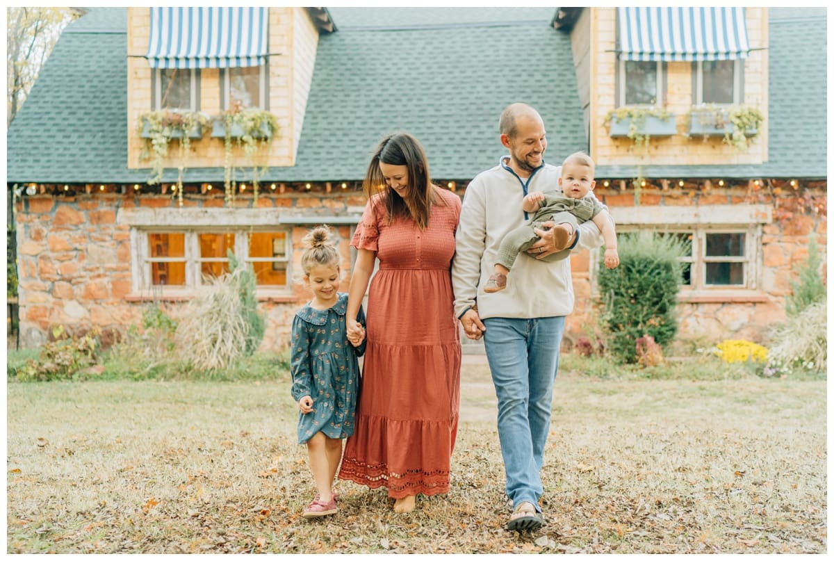 Edmond family of four walking hand in hand in front of a rustic stone house, mom in a long rust dress and dad in casual fall layers holding their baby boy.