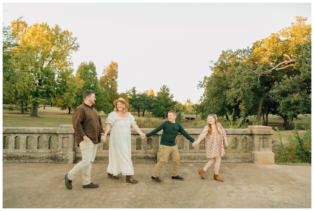 Family of four holds hands while walking across a stone bridge in Edmond. Mom in a white dress and hat, dad in a brown shirt, son in green sweater, and daughter in a floral dress.