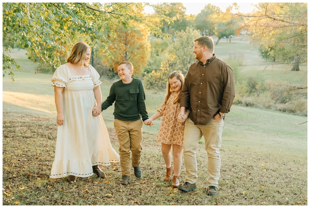 Parents and two children walk hand in hand through a grassy park at golden hour, laughing together in coordinated fall outfits during Edmond family portraits.