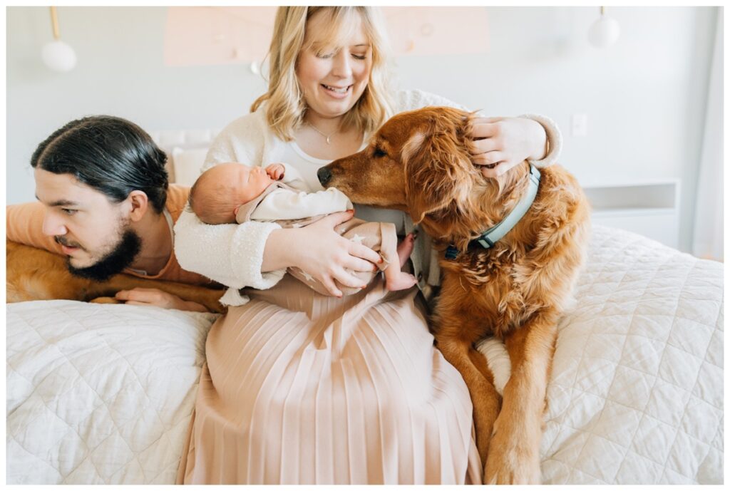 mom and dad on edge of bed with newborn baby and family dog