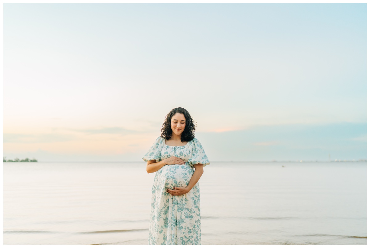 Scenic maternity photo of a pregnant mom holding her baby bump in front of a calm lake at sunset, with soft golden light and peaceful water in the background.