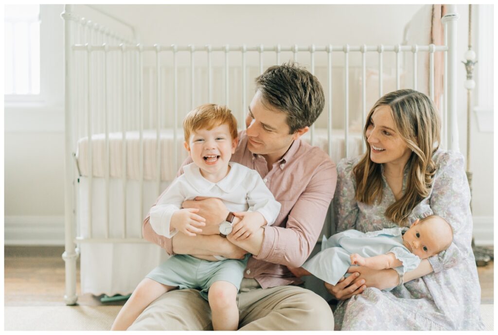 family of four sitting infront of crib with toddler and newborn in laps