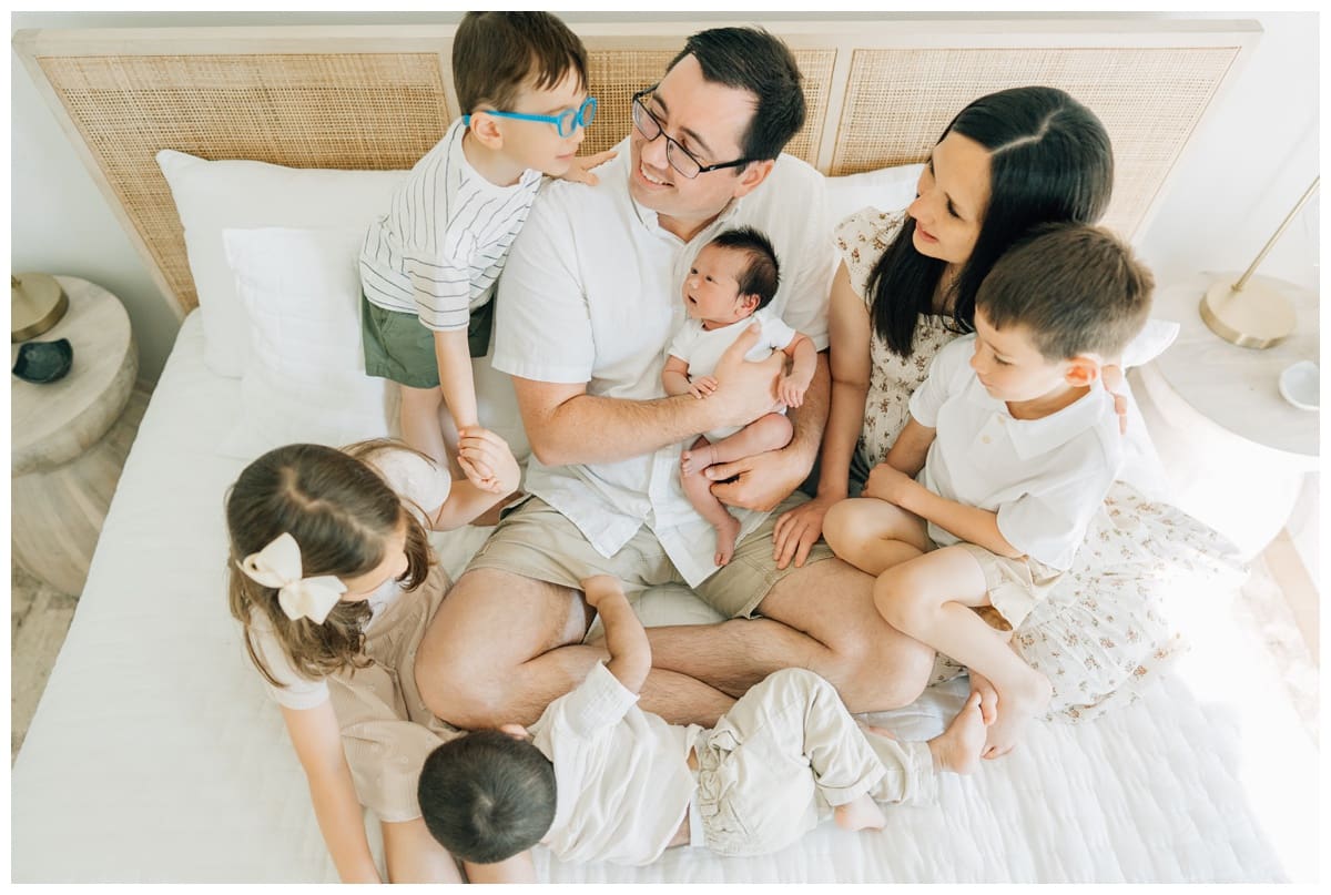 Family poses on the bed with newborn in the father’s arms, everyone smiling together.