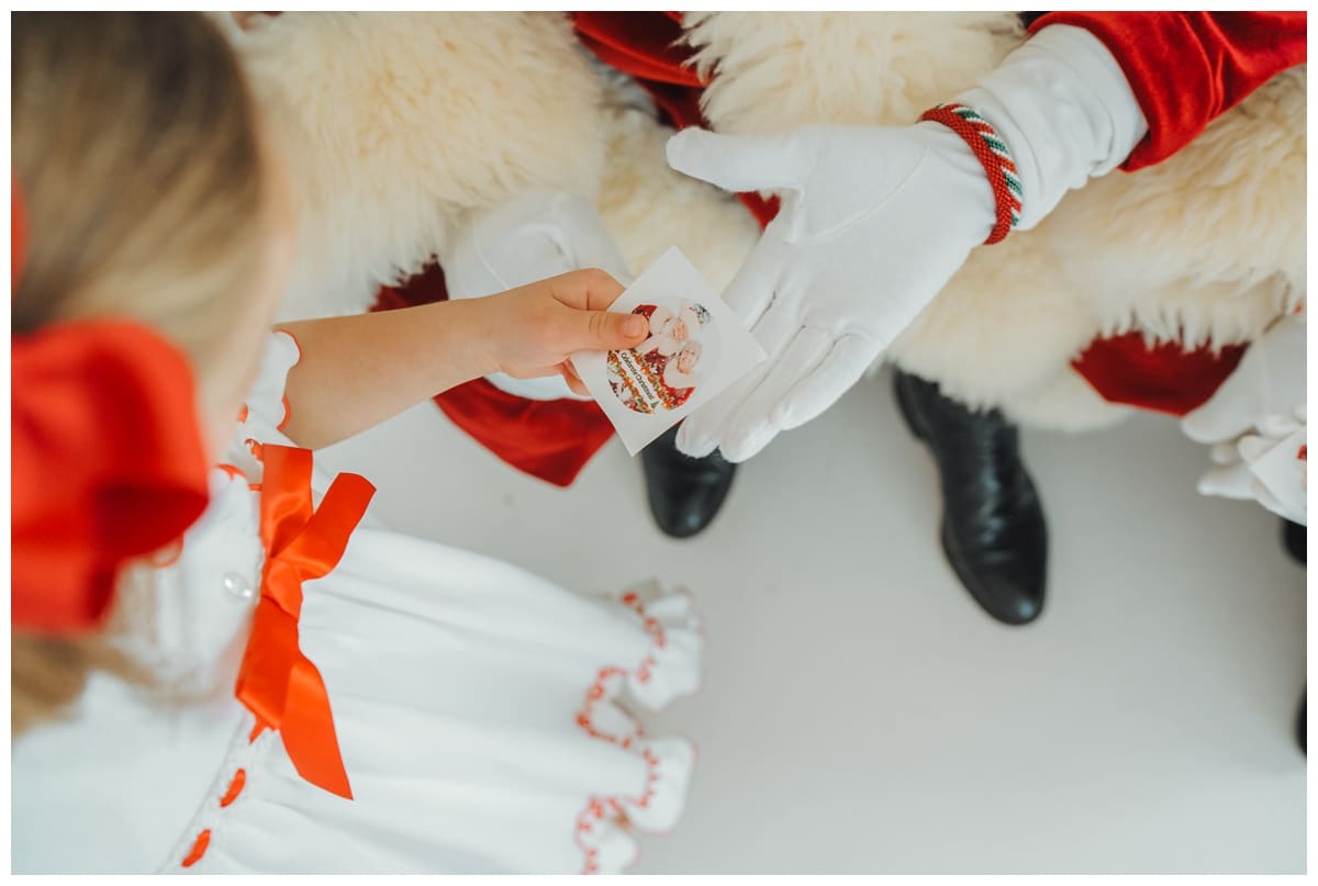 Child in a white dress with red bows hands Santa a keepsake photo card during a sweet Santa Minis holiday session.