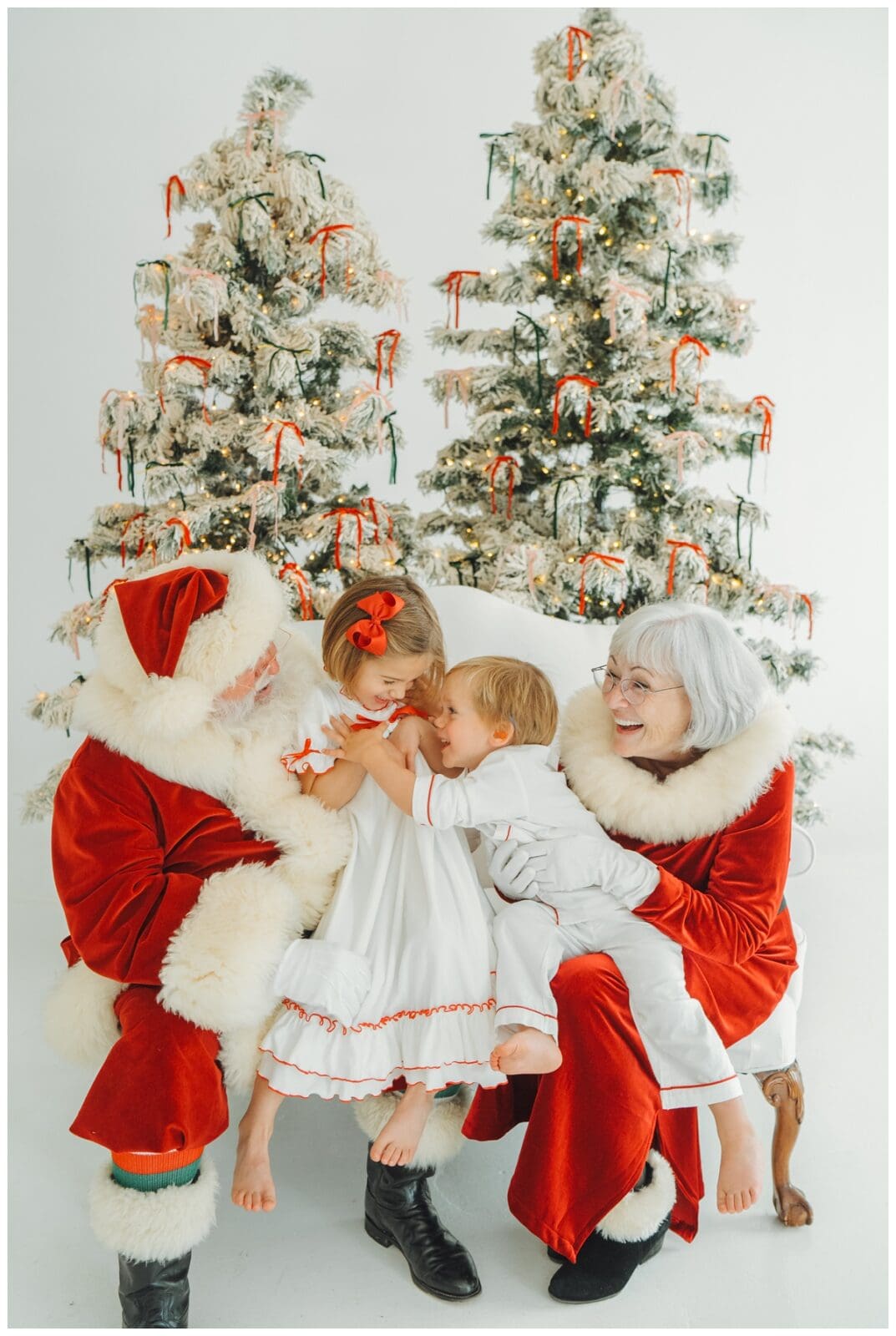 Santa and Mrs. Claus hug two siblings in matching white and red holiday pajamas during joyful Santa Minis portraits.